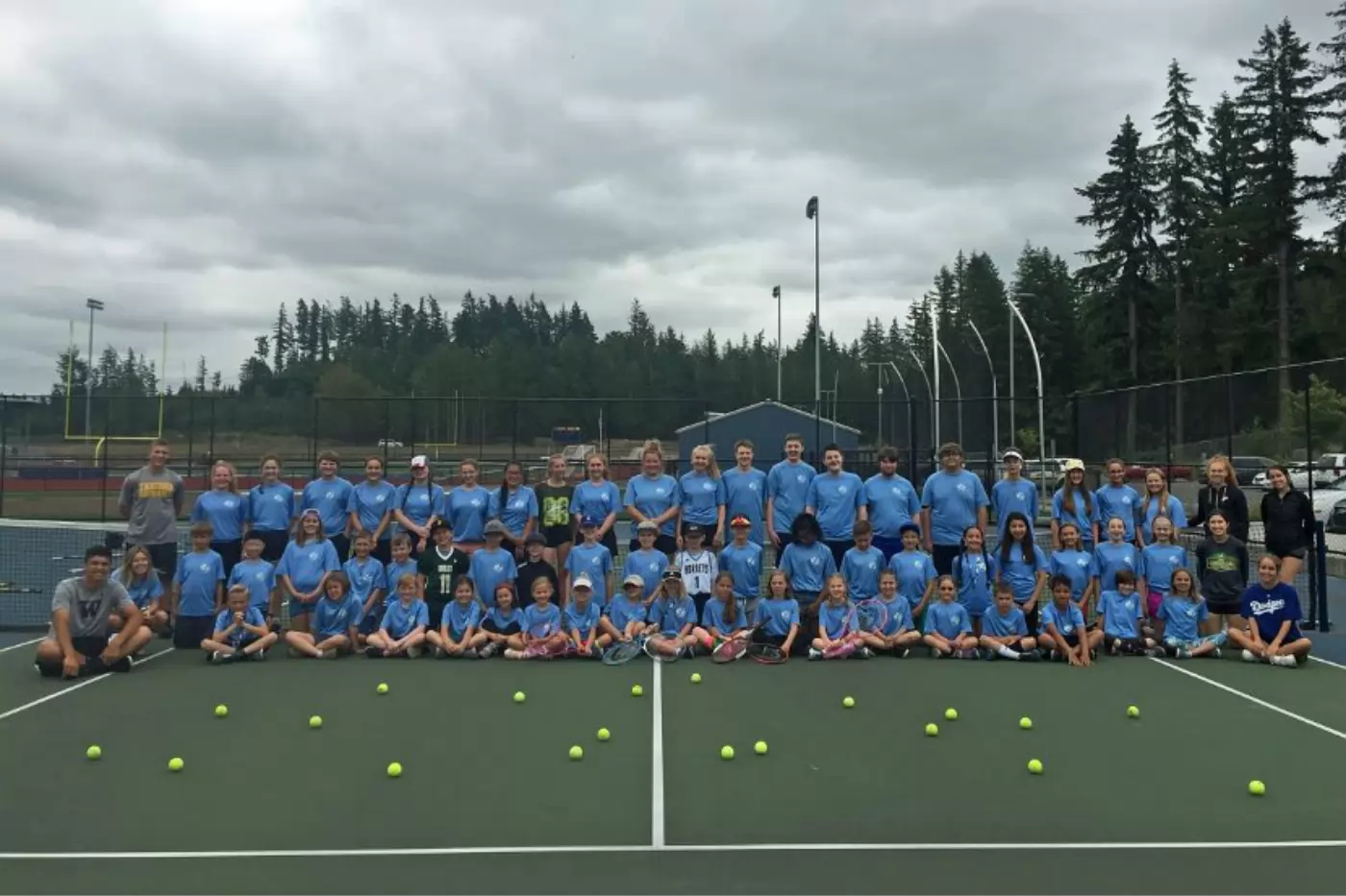 Group photo of children and adults in blue shirts posing on a tennis court, with tennis balls on the ground. Trees and a sports field are visible in the background.