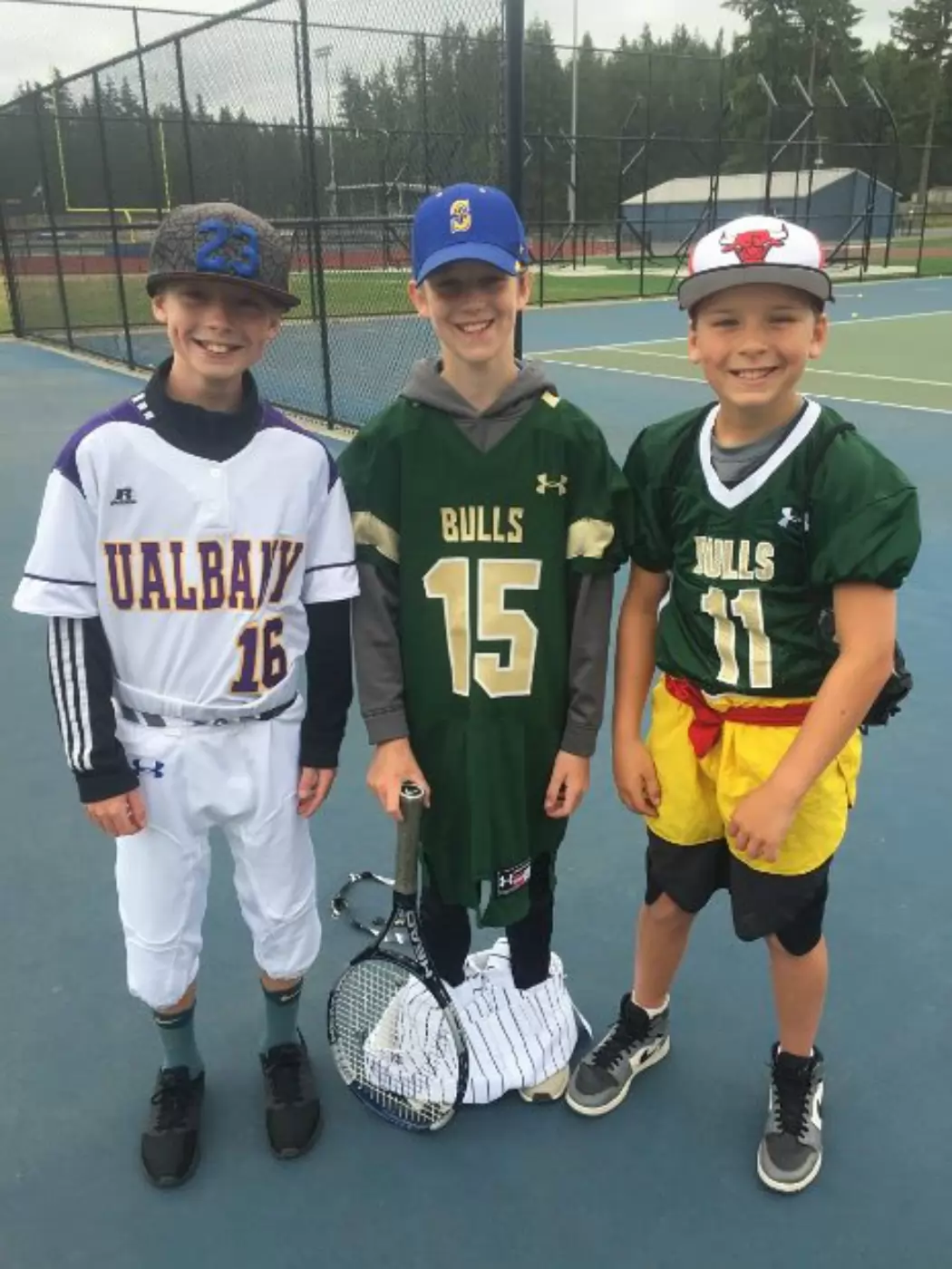 Three boys wearing sports jerseys and caps stand together on a tennis court, smiling. One holds a tennis racket, and the others have football jerseys. A field is visible behind them.