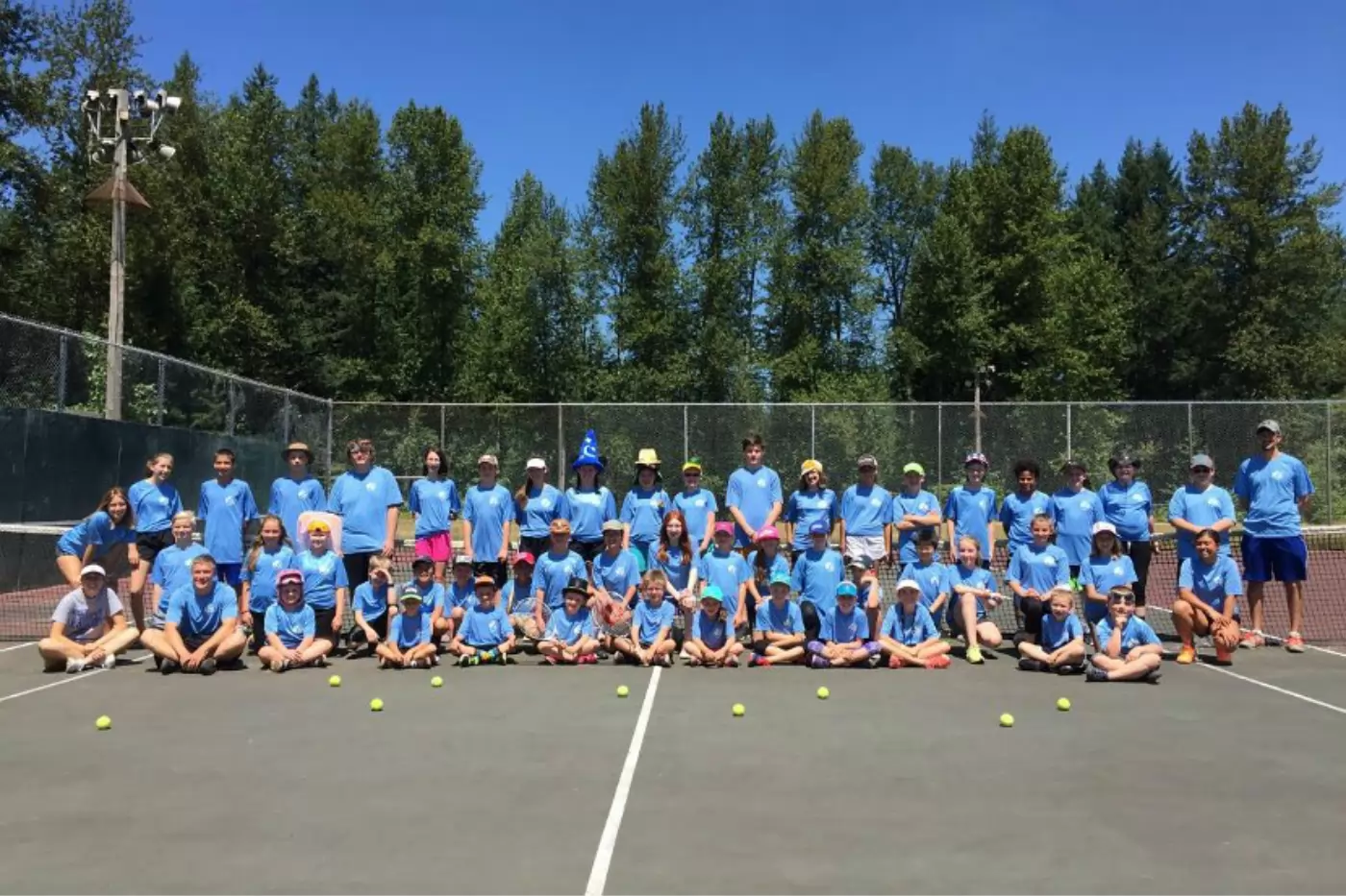 A large group of people wearing matching blue shirts and hats posing on a tennis court with tennis balls. The background features tall trees and a clear blue sky.