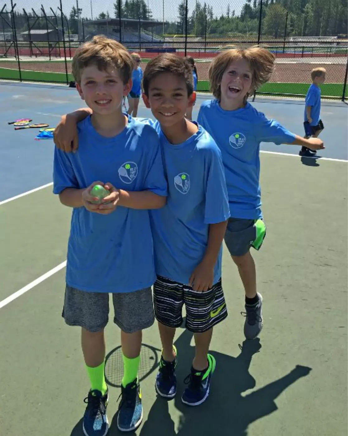 Three boys in matching blue sports shirts smile on a tennis court. One boy holds a green ball, while another poses playfully behind. Tennis rackets are on the ground.
