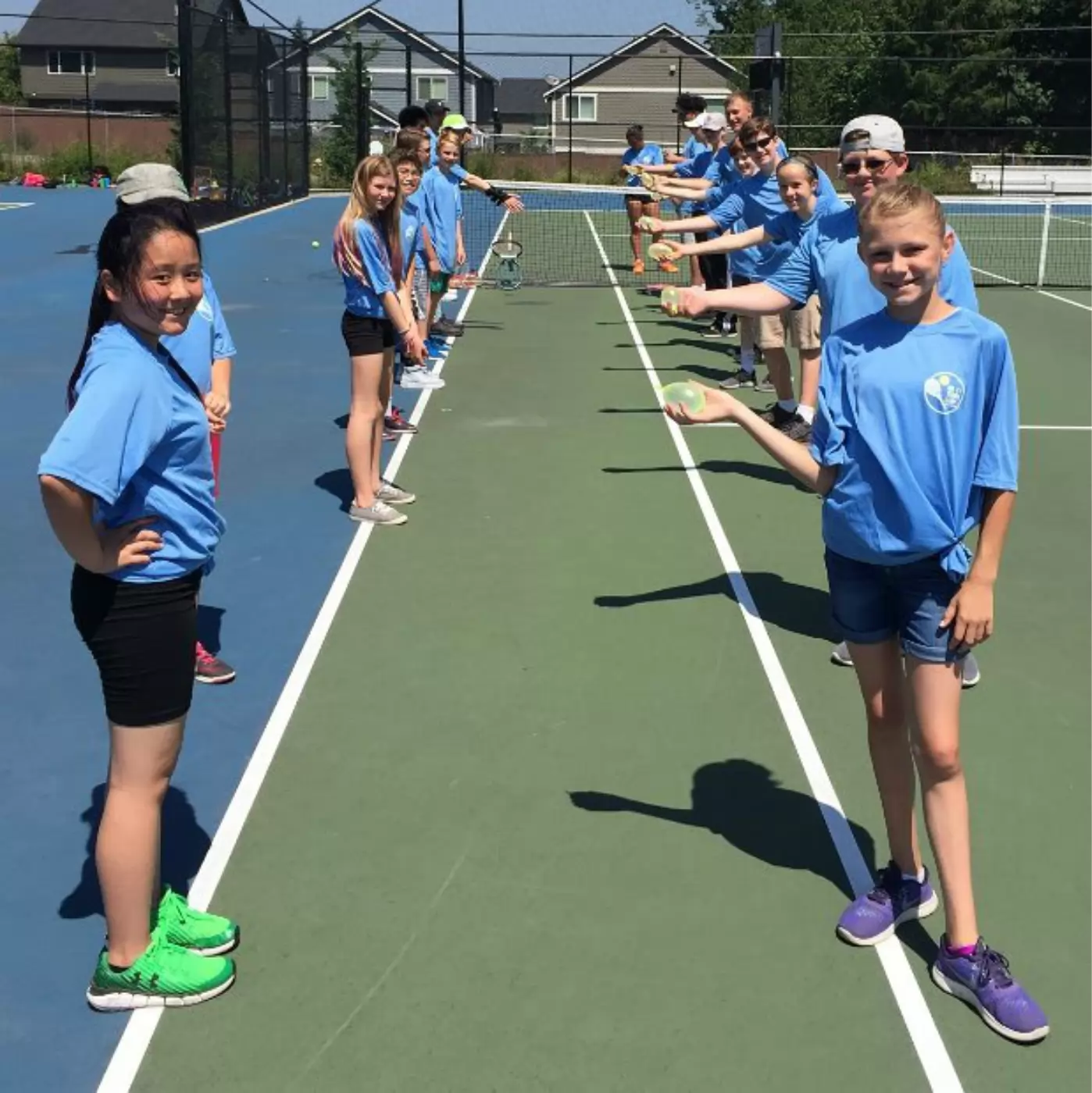 A group of kids on a tennis court, wearing matching blue shirts. They stand in two parallel lines, each holding a water balloon, smiling and facing the camera.