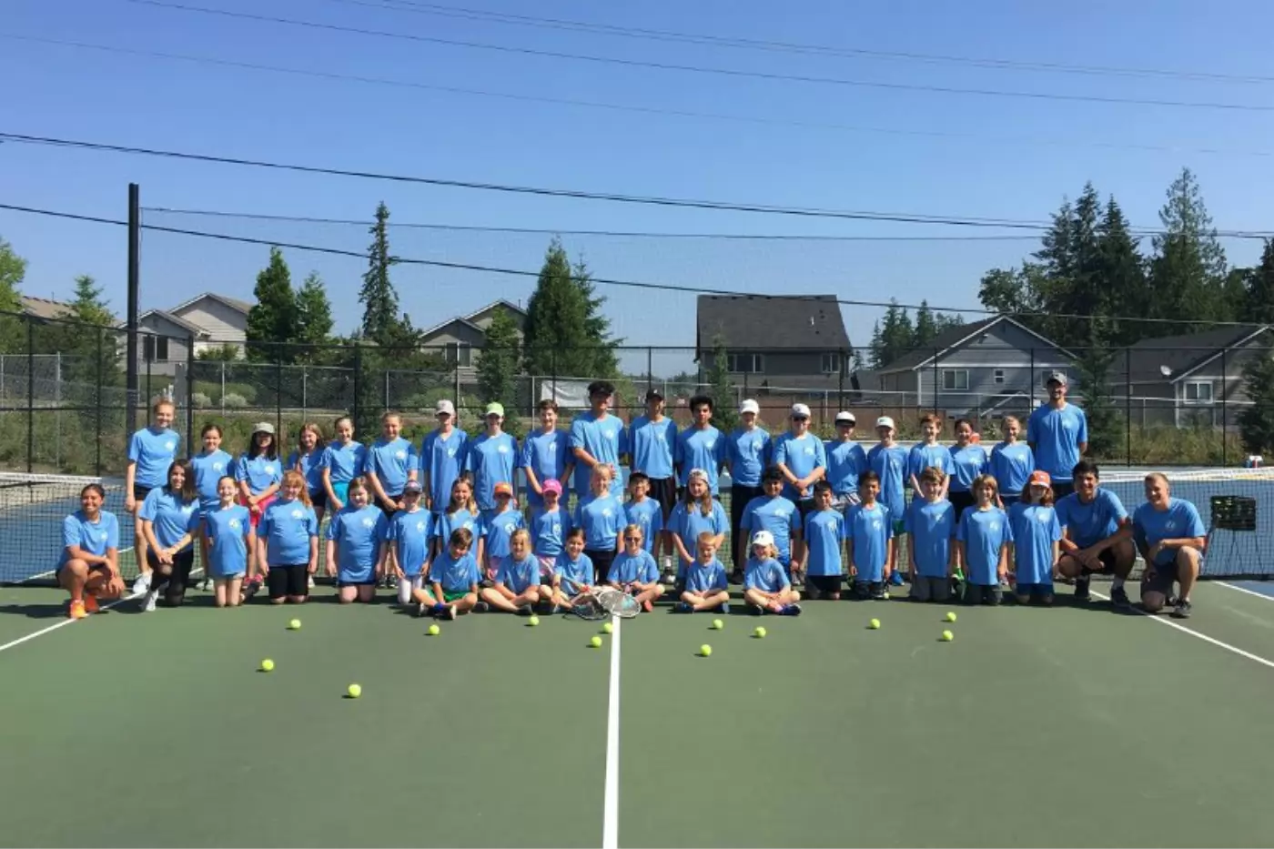 Group of people in matching blue shirts on a tennis court, posing for a photo. Tennis balls are scattered on the ground in front of them. Houses and trees are in the background.