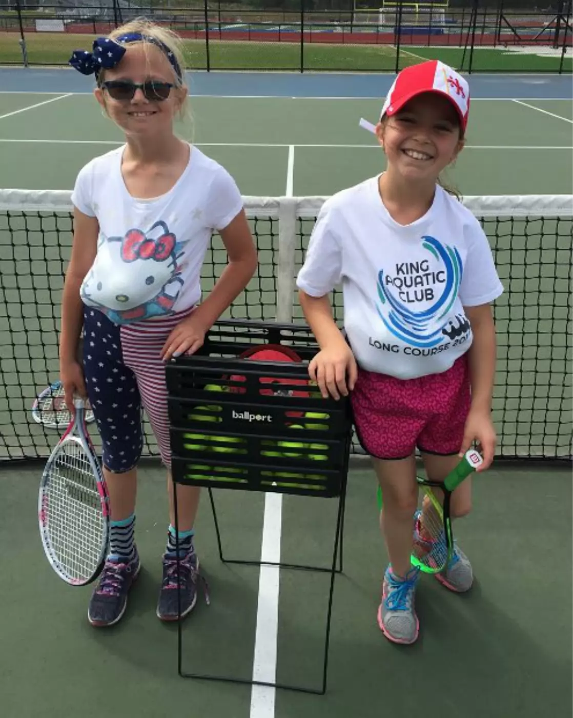 Two young girls stand on a tennis court smiling. One wears a bow, sunglasses, and a Hello Kitty shirt; the other wears a red cap and 