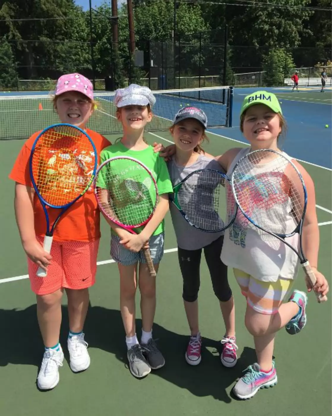 Four children on a tennis court pose with rackets, smiling. They are dressed in colorful summer clothes and hats under a sunny sky, with tennis nets and trees in the background.