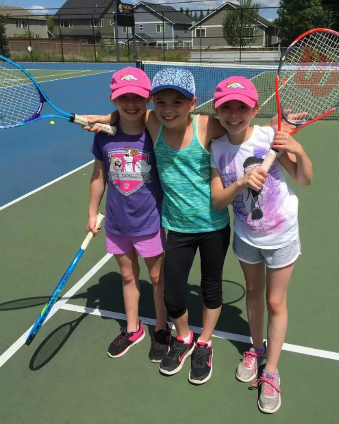 Three young girls smiling on a tennis court, each holding a tennis racket. They wear colorful caps and casual athletic outfits, standing together with arms around each other.