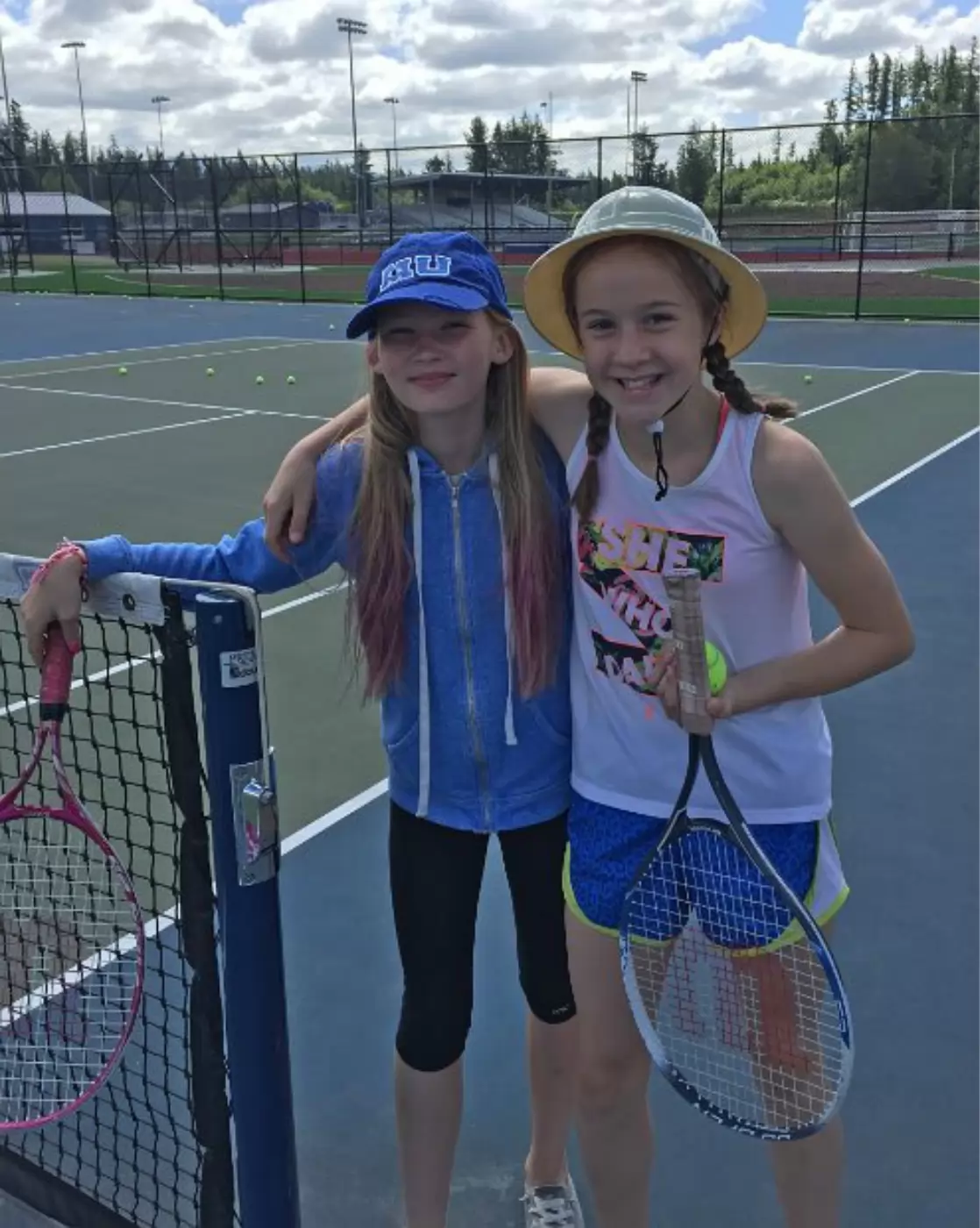 Two young girls smiling on a tennis court. One wears a blue hoodie and cap, the other in a tank top and bucket hat. Both hold tennis rackets next to a net.