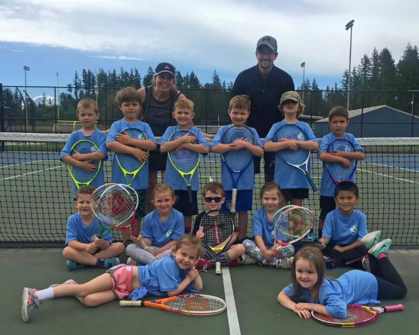 Group of kids in blue shirts pose with tennis rackets on a court, flanked by two adults. Some children kneel in front, while the rest stand in a row behind them.