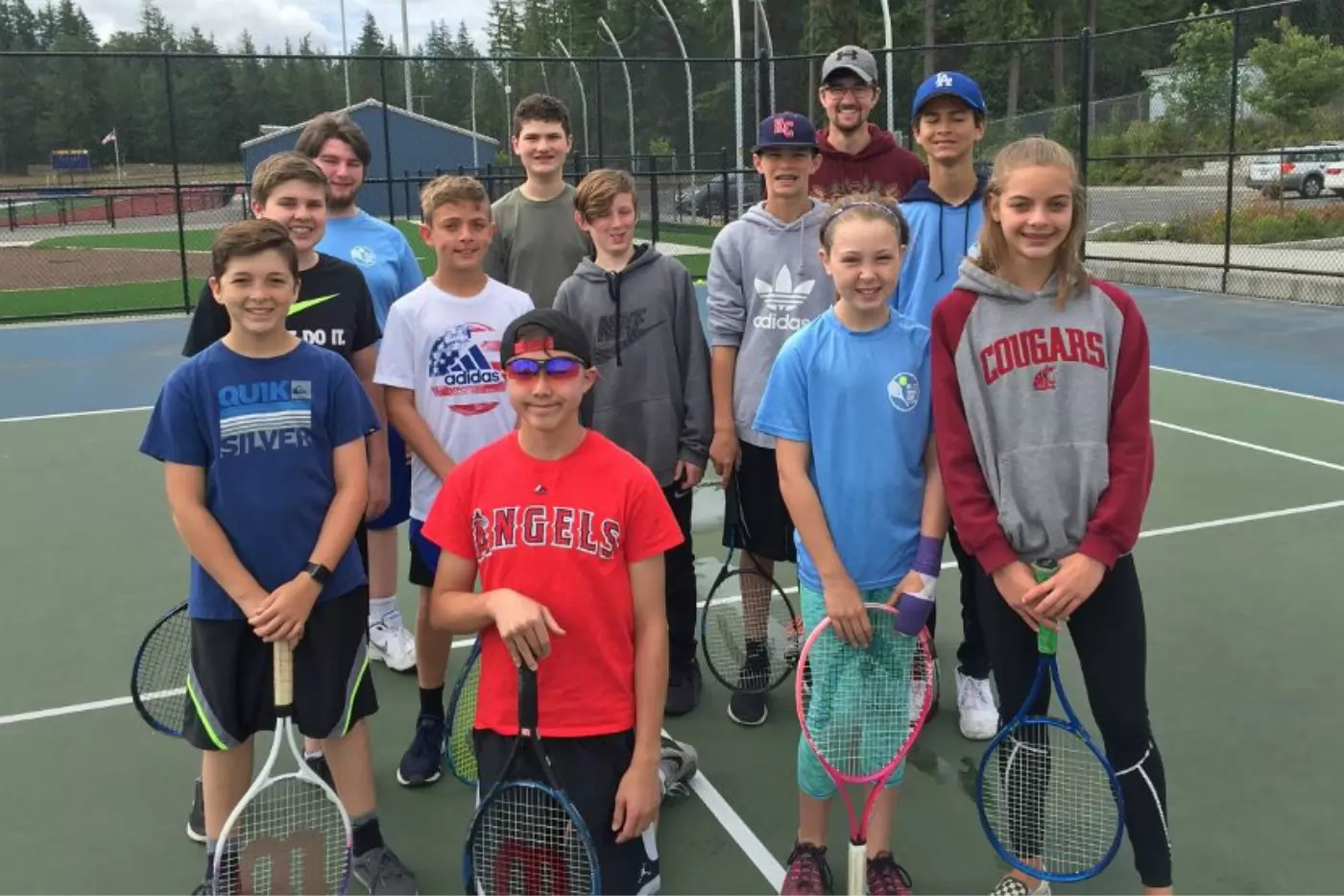 A group of young people holding tennis rackets stands on a tennis court, posing for a photo. There are trees and a small building in the background.