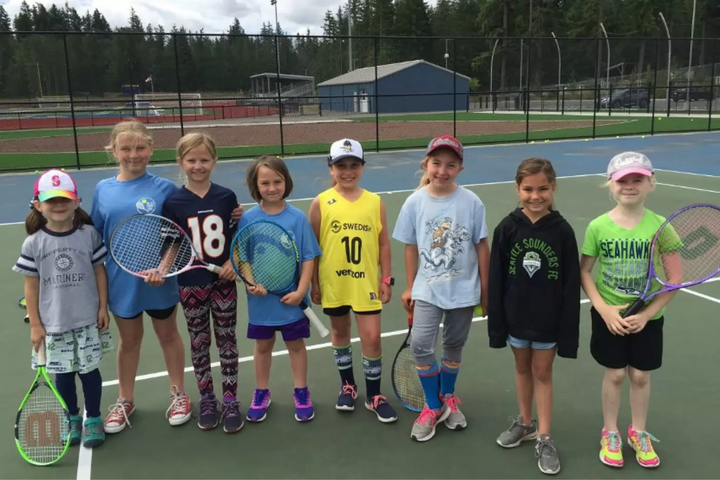 Nine children holding tennis rackets stand in a row on an outdoor tennis court, smiling at the camera. They are dressed in casual sports attire. Trees and a building are in the background.