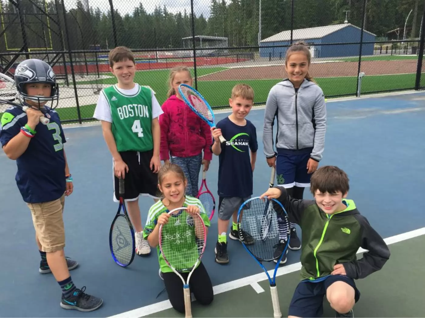 A group of seven children holding tennis rackets stand on a court, dressed in sports attire, including jerseys and a football helmet, with a green field and trees in the background.