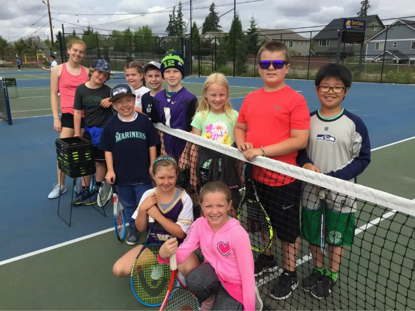 A group of children and one adult standing and kneeling on a tennis court, holding rackets, near a net, smiling. The background has trees and suburban houses.