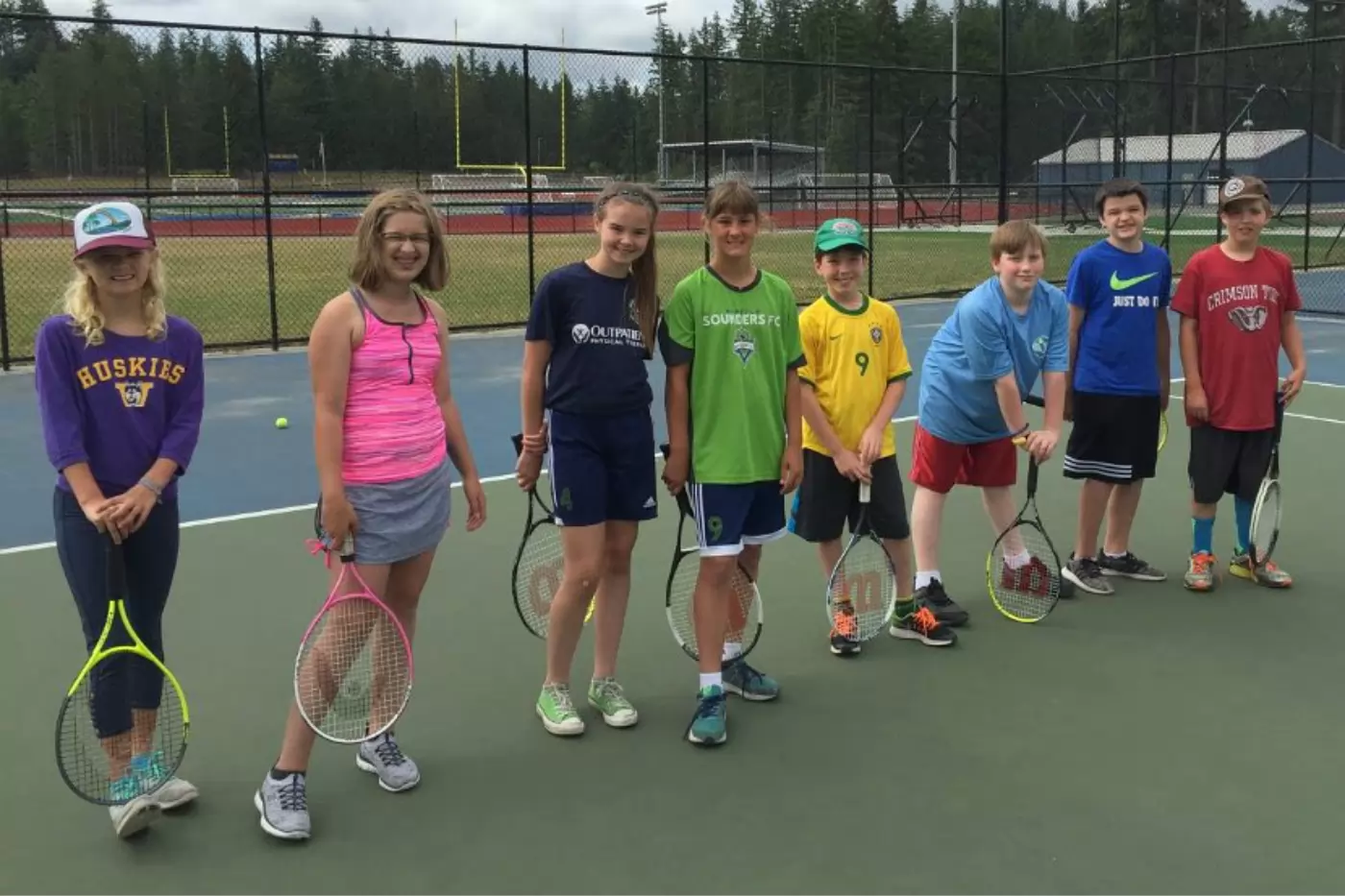 A group of eight kids standing on a tennis court, holding rackets. They are dressed casually in various sports shirts and caps, with a chain-link fence and trees in the background.