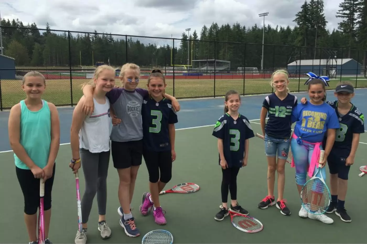 A group of eight children stands on a tennis court holding rackets, smiling. They wear casual athletic clothing and jerseys. A sports field and trees are visible in the background.