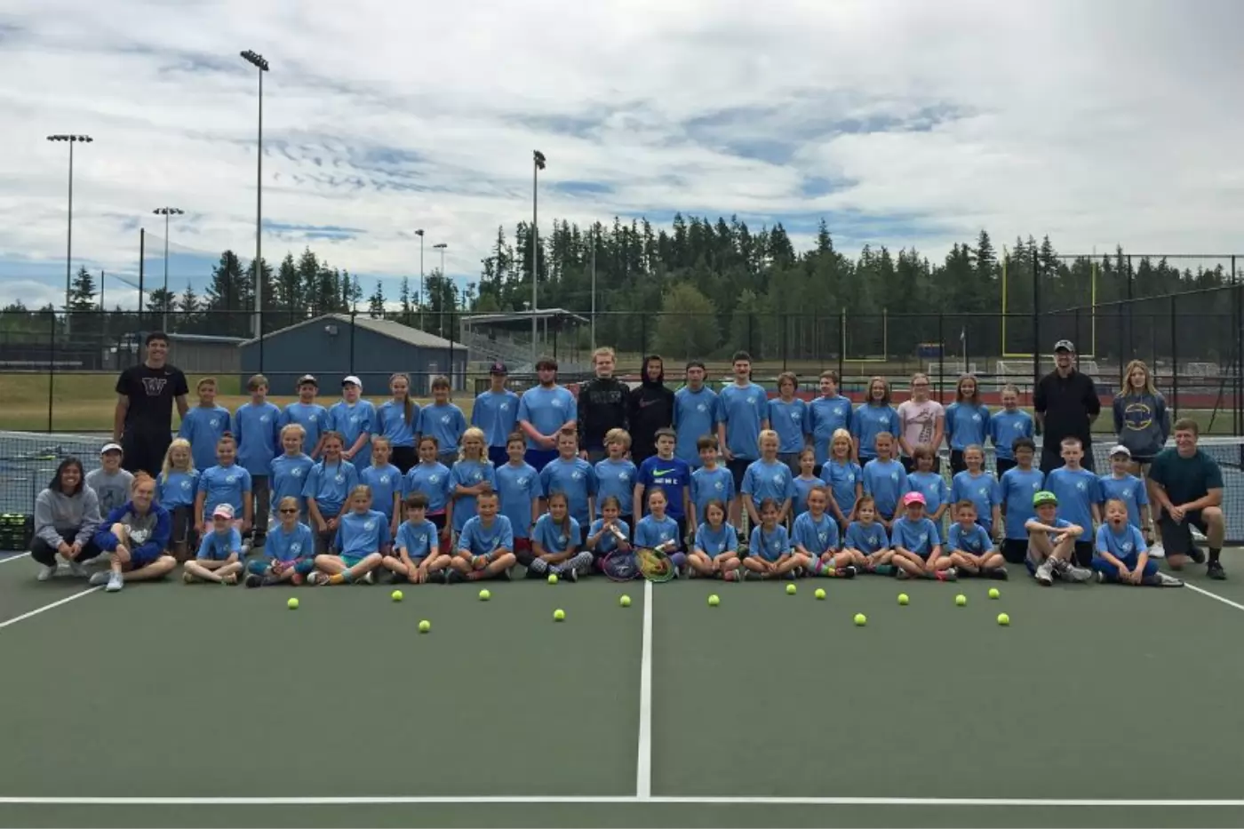 Group of children and adults in matching blue shirts standing on a tennis court, with tennis balls lined up in front of them. Trees and sports fields visible in the background.