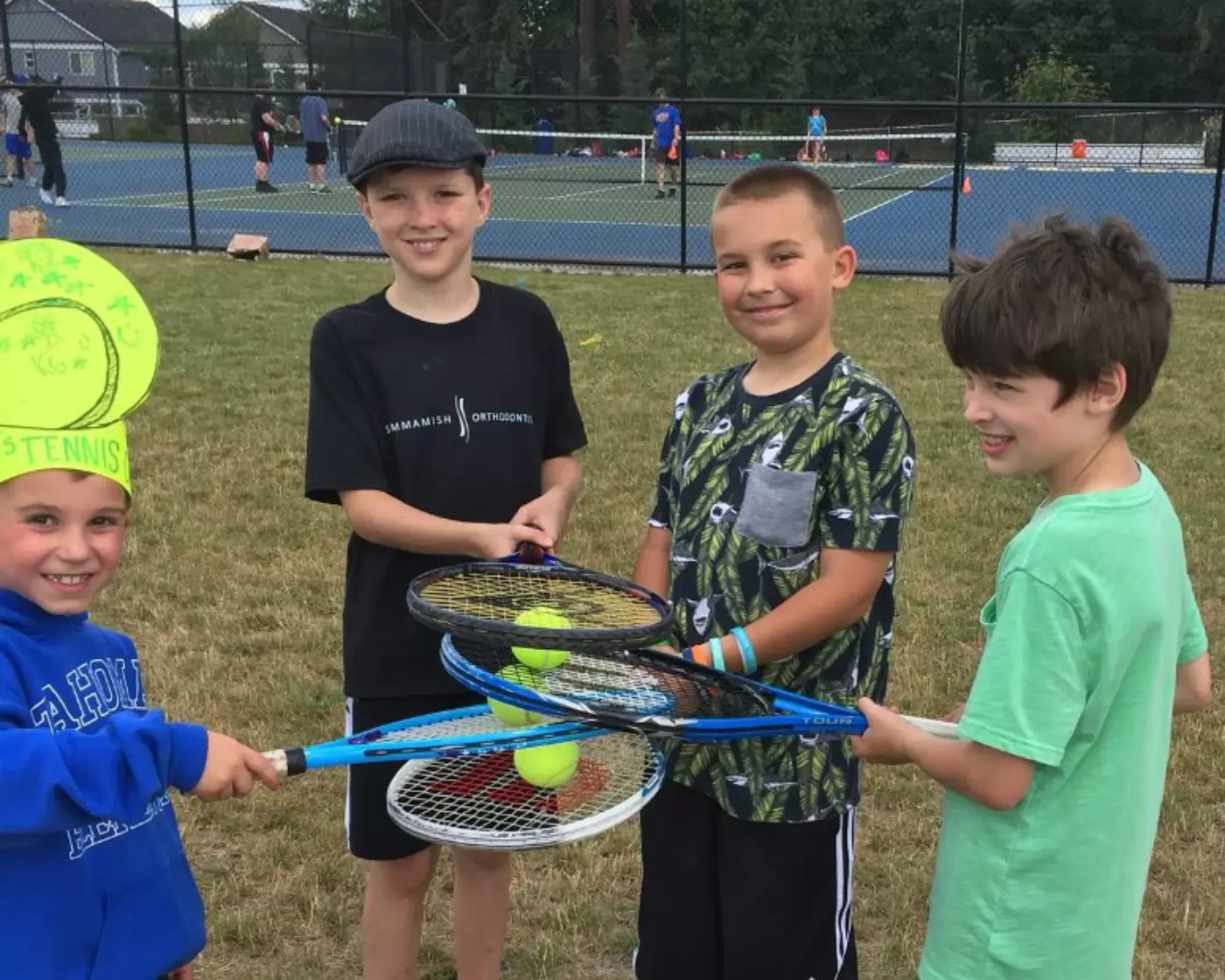 Four boys are standing on a grass field smiling, holding tennis rackets piled with tennis balls. One wears a bright yellow paper hat. Tennis courts are in the background.