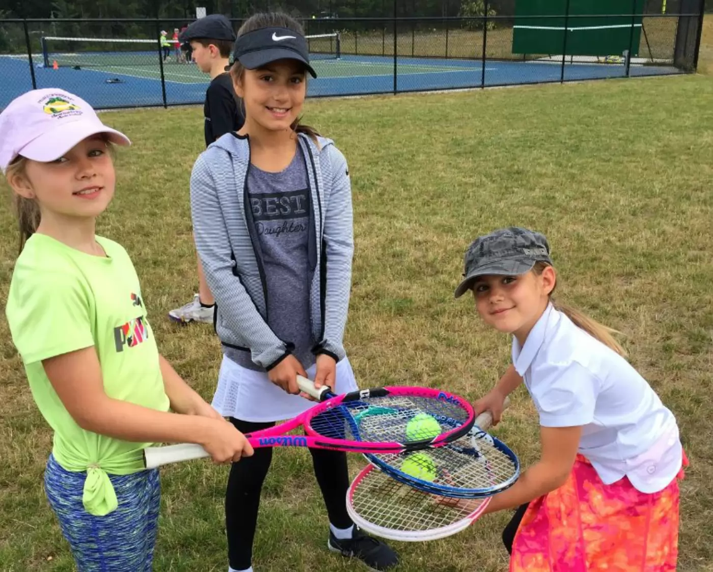 Three young girls in sportswear and caps hold tennis rackets with tennis balls on them, smiling on a grassy field near a tennis court. A boy stands in the background.