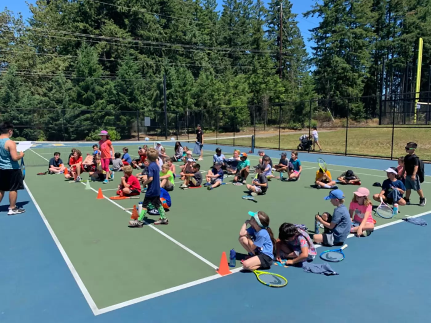 A group of children sitting on an outdoor tennis court, holding rackets and listening to an adult instructor. The court is surrounded by trees under a clear blue sky.