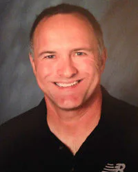 A smiling man with short hair wearing a dark polo shirt stands against a soft-focus blue background.