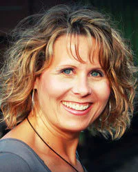 Smiling woman with curly blonde hair wearing a gray top and hoop earrings, standing outdoors.