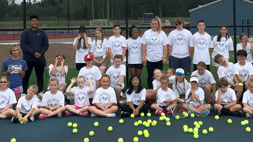 A group of children and adults wearing matching white tennis shirts pose in front of a fence on a tennis court, surrounded by scattered yellow tennis balls.