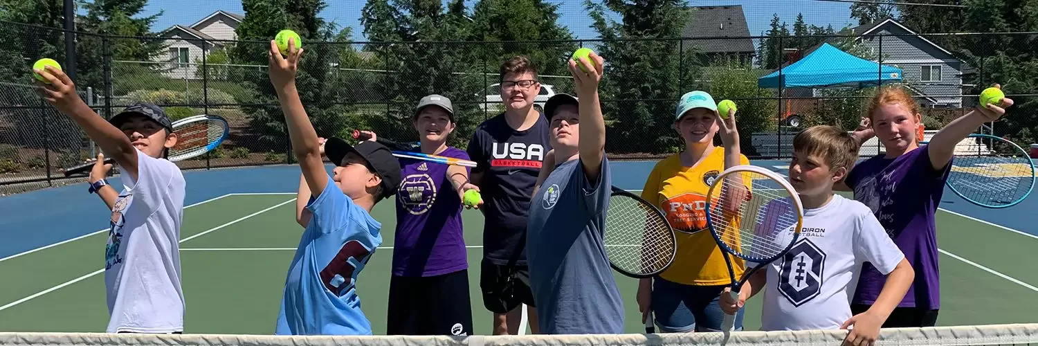 A group of kids and a coach on an outdoor tennis court, each holding tennis rackets and tennis balls, posing energetically in a sunny setting with trees and houses in the background.