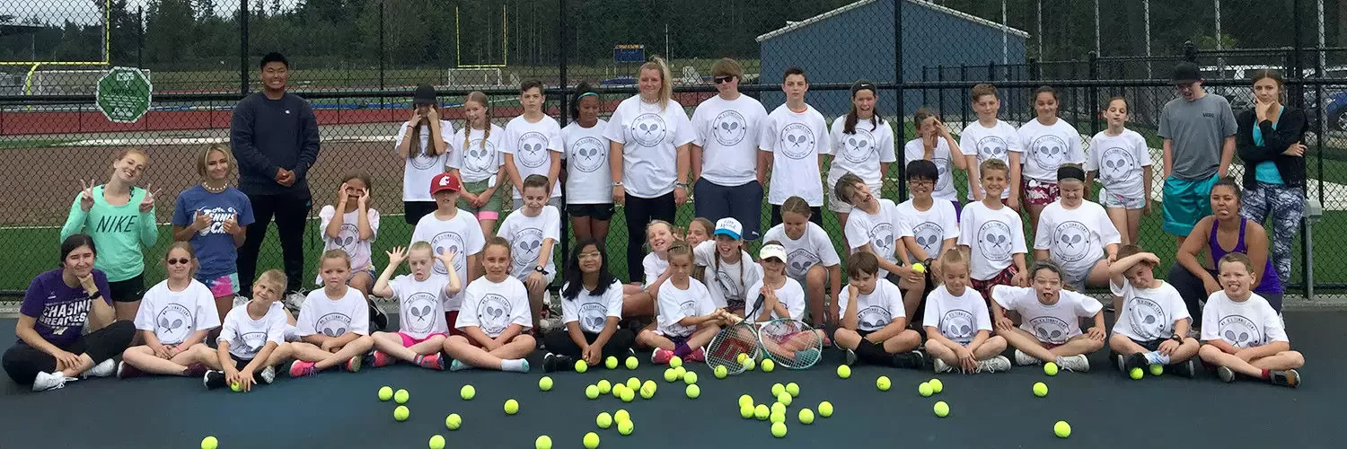 A group of children and adults wearing matching white tennis shirts pose in front of a fence on a tennis court, surrounded by scattered yellow tennis balls.
