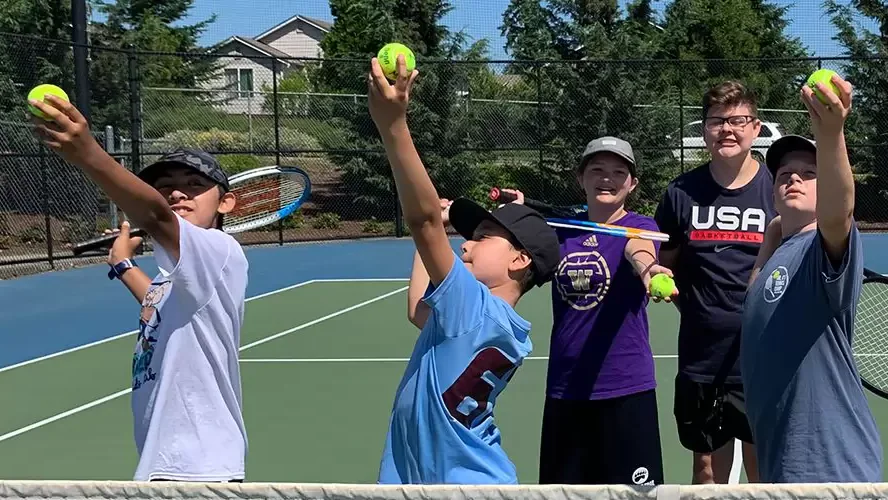 A group of kids and a coach on an outdoor tennis court, each holding tennis rackets and tennis balls, posing energetically in a sunny setting with trees and houses in the background.
