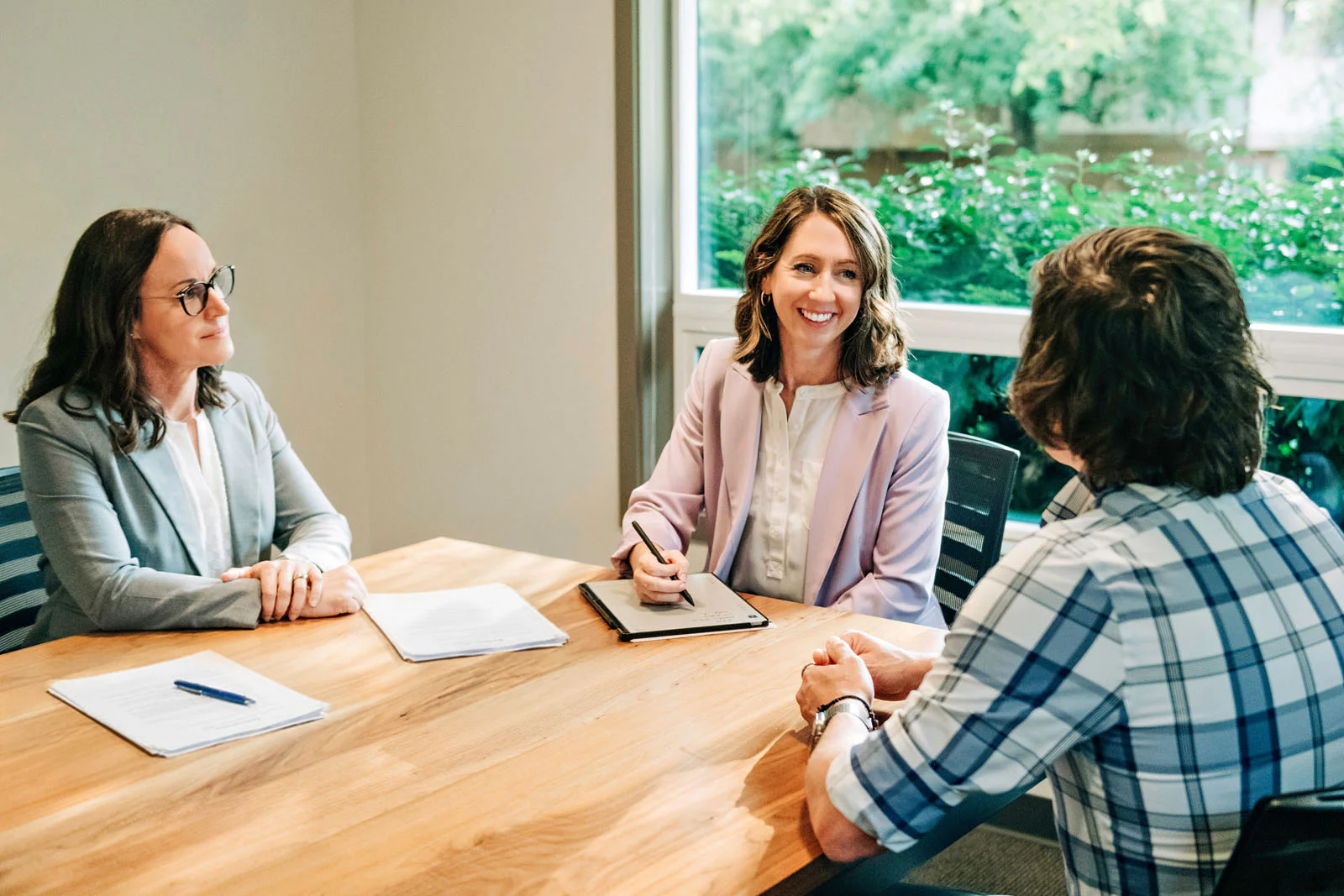 Three people are sitting at a table in a meeting room. Two women are on one side with papers, and a man is on the other side. One woman is smiling and taking notes.