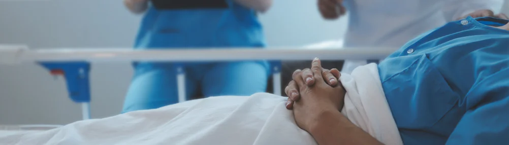 Patient lying in a hospital bed with hands clasped, wearing a blue gown. Medical staff in blue scrubs stand nearby, partially visible in the background.