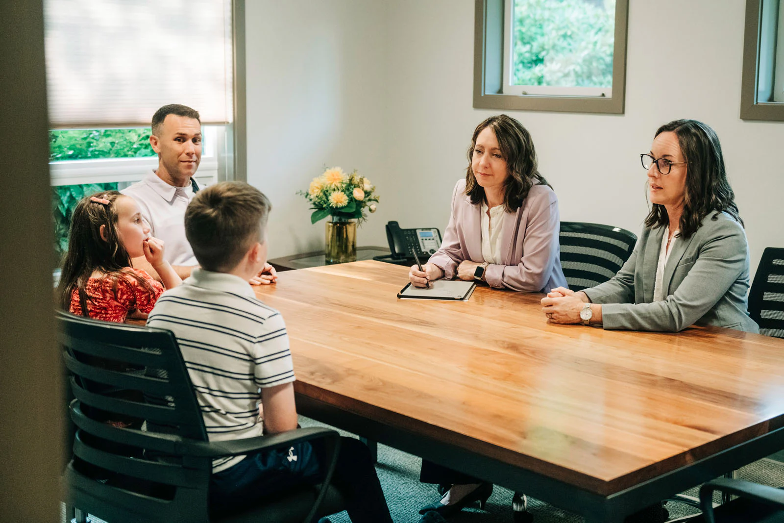 A group of five people, including two children and three adults, sits around a wooden conference table in a meeting room with flowers on a table and a phone in the background.