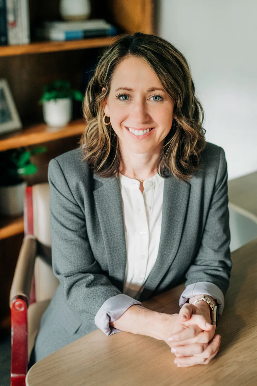 Smiling woman in a gray blazer and white blouse sits at a wooden desk in an office setting, with bookshelves and plants in the background. Her hands are clasped in front.
