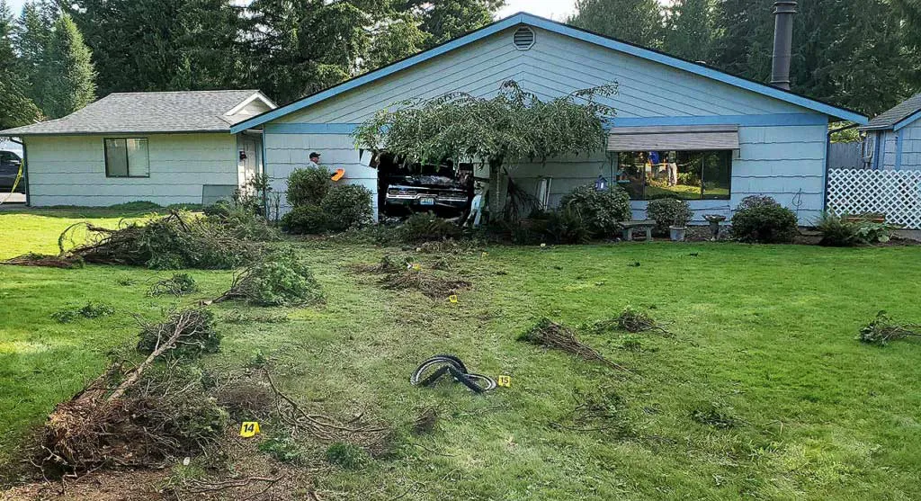 Car crashed into a blue house, damaging the front wall. Debris and tire tracks are visible on the lawn. A person in an orange vest is examining the scene.