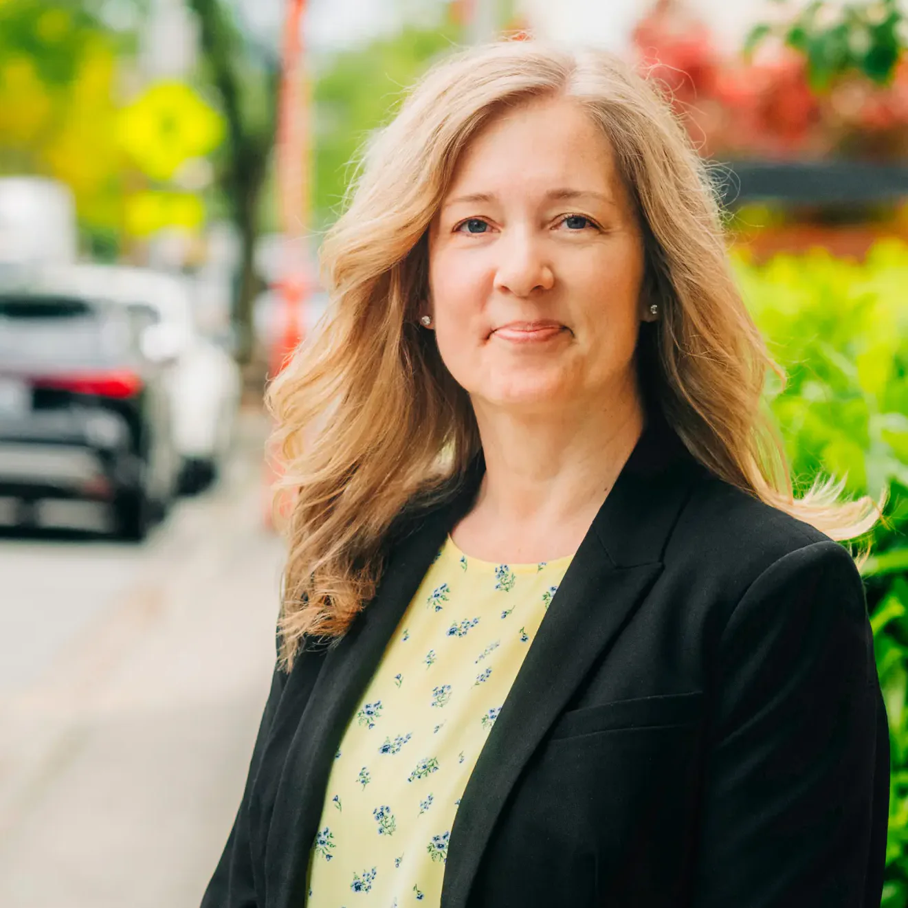 Woman in a black blazer and floral top standing on a sidewalk by green bushes, smiling at the camera