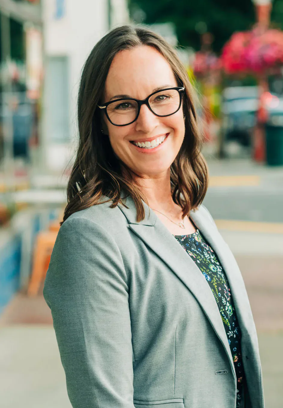 Smiling woman wearing glasses and a light gray blazer stands outdoors on a city street, with blurred background of shops and flowers.