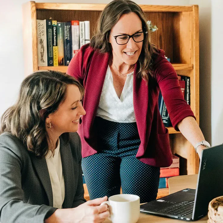 Two women in business attire smiling and discussing something on a laptop screen at a wooden table. One is seated with a mug, while the other stands, pointing at the laptop.