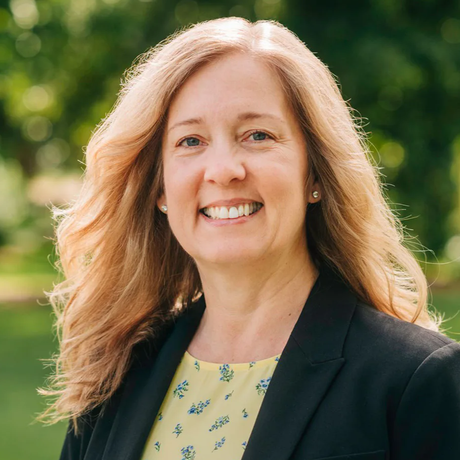Smiling middle-aged woman with long blonde hair in a black blazer and yellow floral blouse outdoors