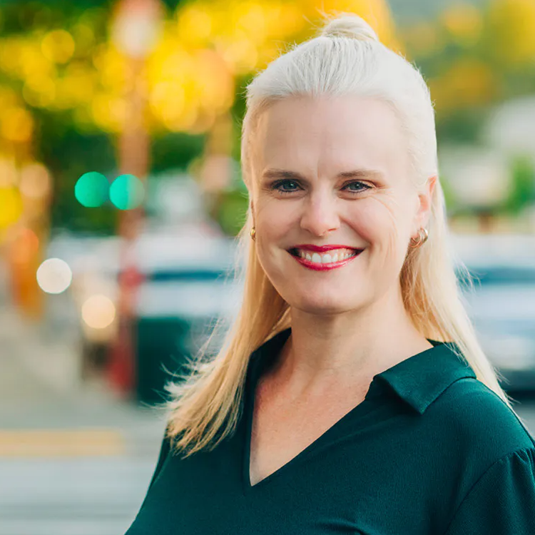 Smiling blonde woman in teal top standing on a city street with blurred traffic and trees behind her.