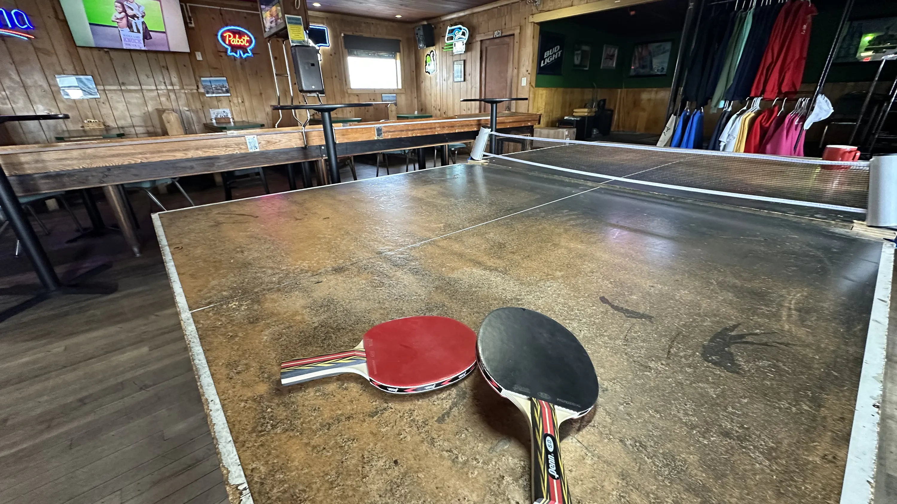 Two table tennis paddles, one red and one black, rest on a ping pong table in a wood-paneled bar with neon signs, a TV, and hanging merch in the background.