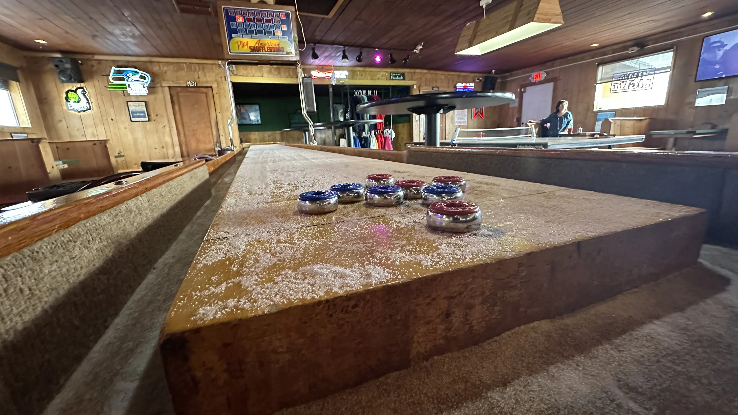 A shuffleboard table with red and blue pucks in a wooden-paneled bar. Neon signs adorn the walls, and a person stands near another table in the background.