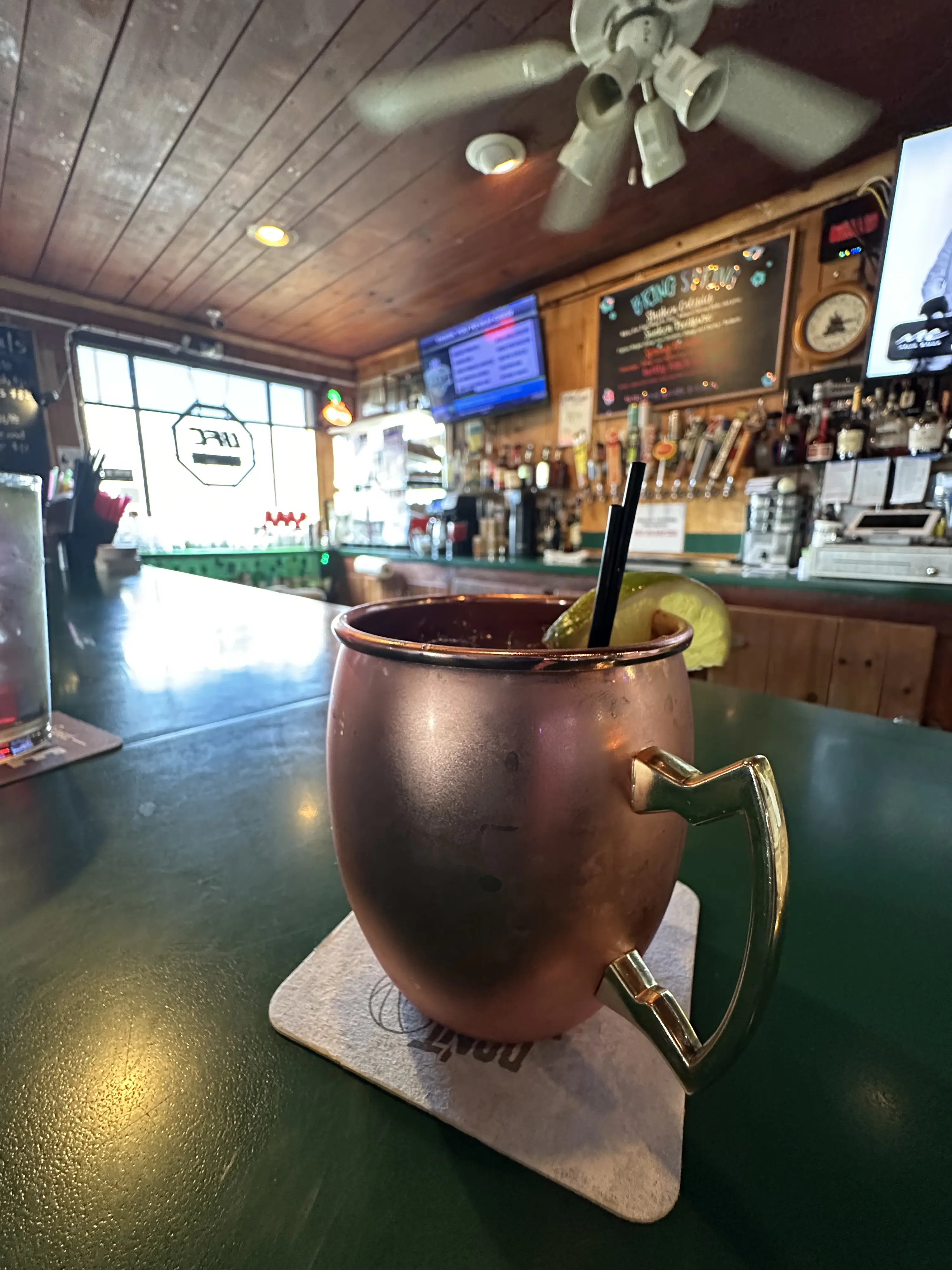 A copper mug with a lime wedge and straw sits on a coaster at a bar with a wooden ceiling, a ceiling fan, TV, and shelves of bottles in the background.