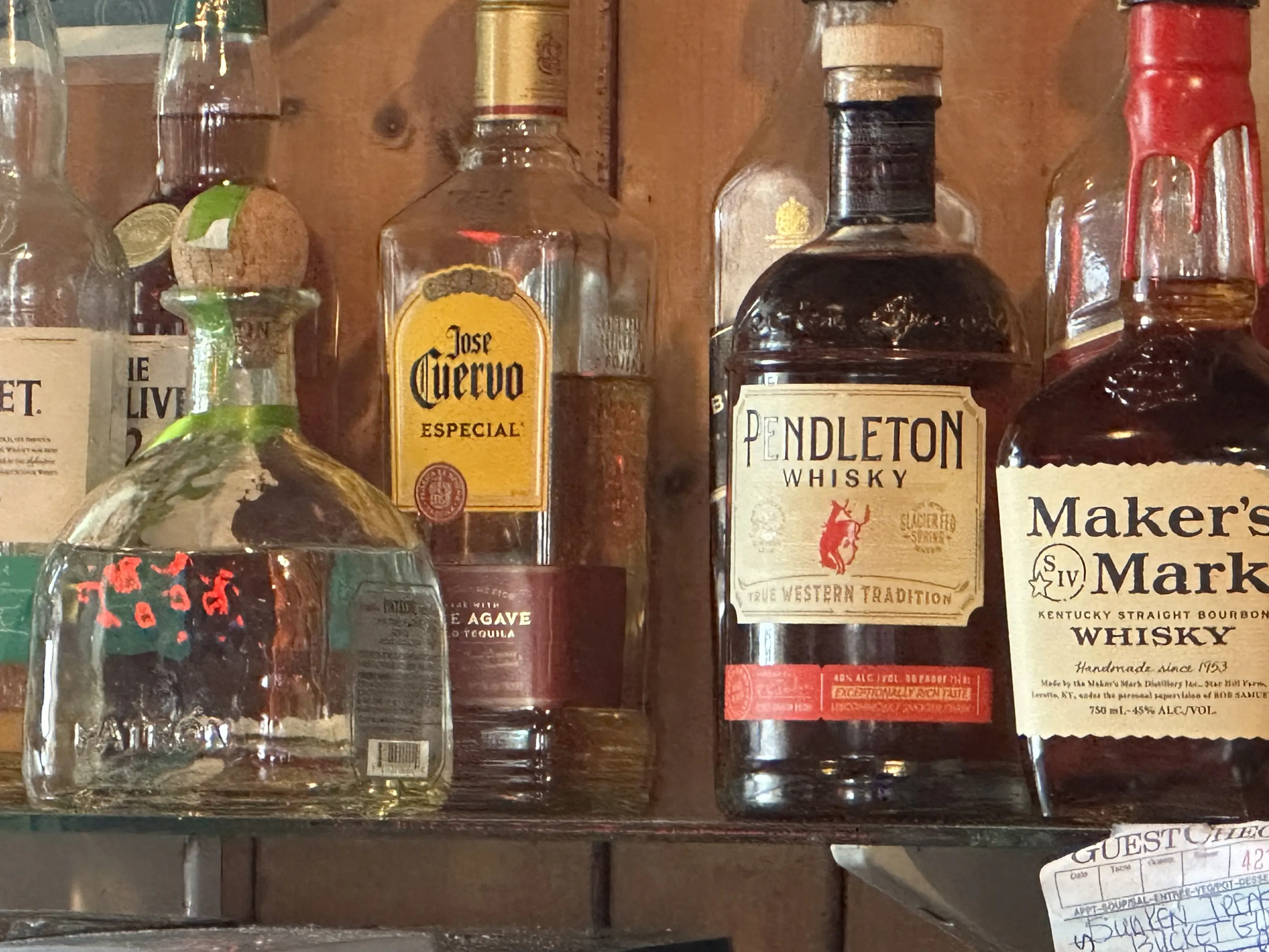 A display of liquor bottles includes brands like Jose Cuervo Especial, Pendleton Whisky, and Maker's Mark. Bottles are arranged on a shelf against a wooden background.