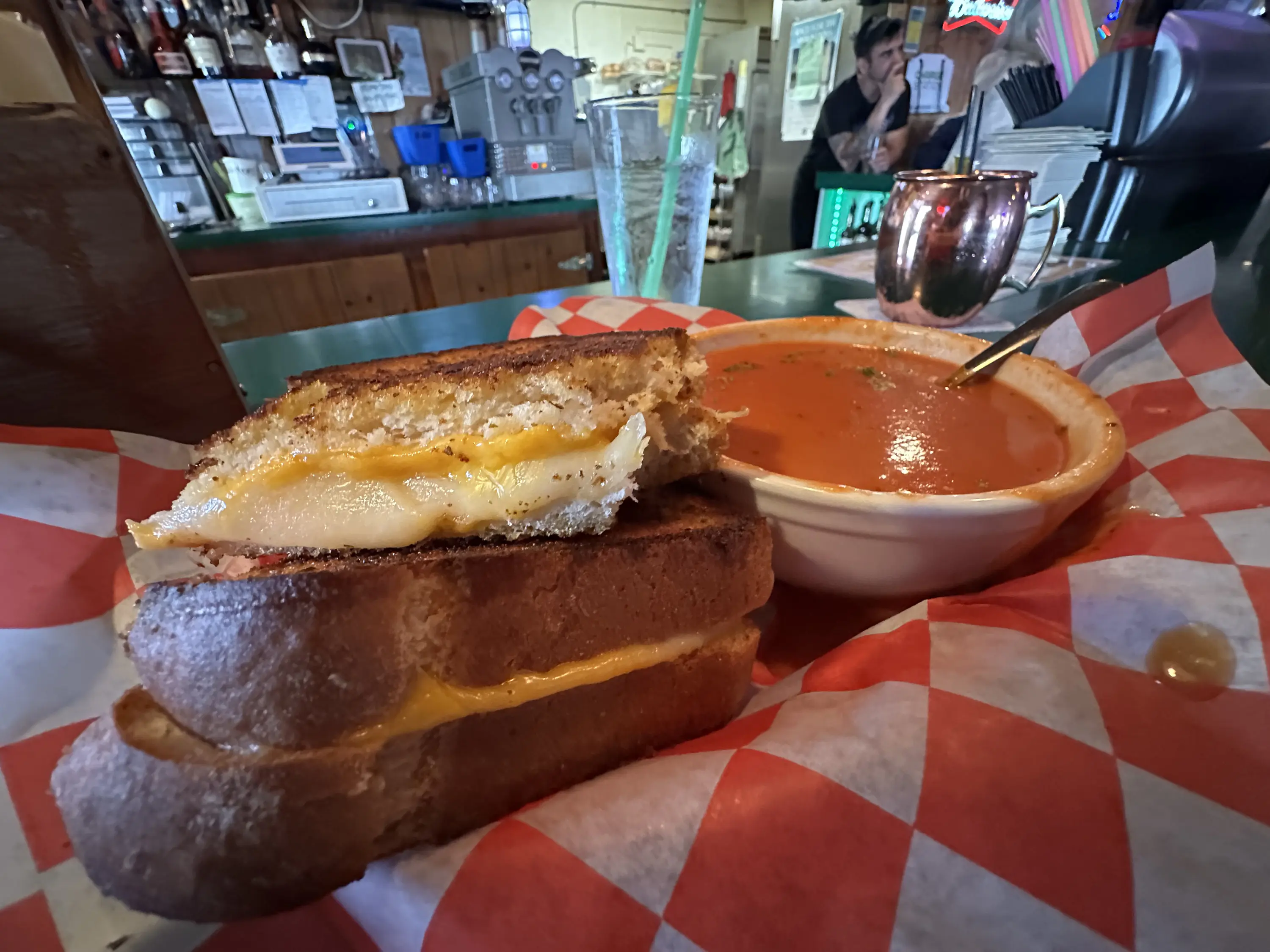 Grilled cheese sandwich with melted cheese on toasted bread, served with a bowl of tomato soup on a red and white checkered paper. Bar in the background with drinks and man sitting.