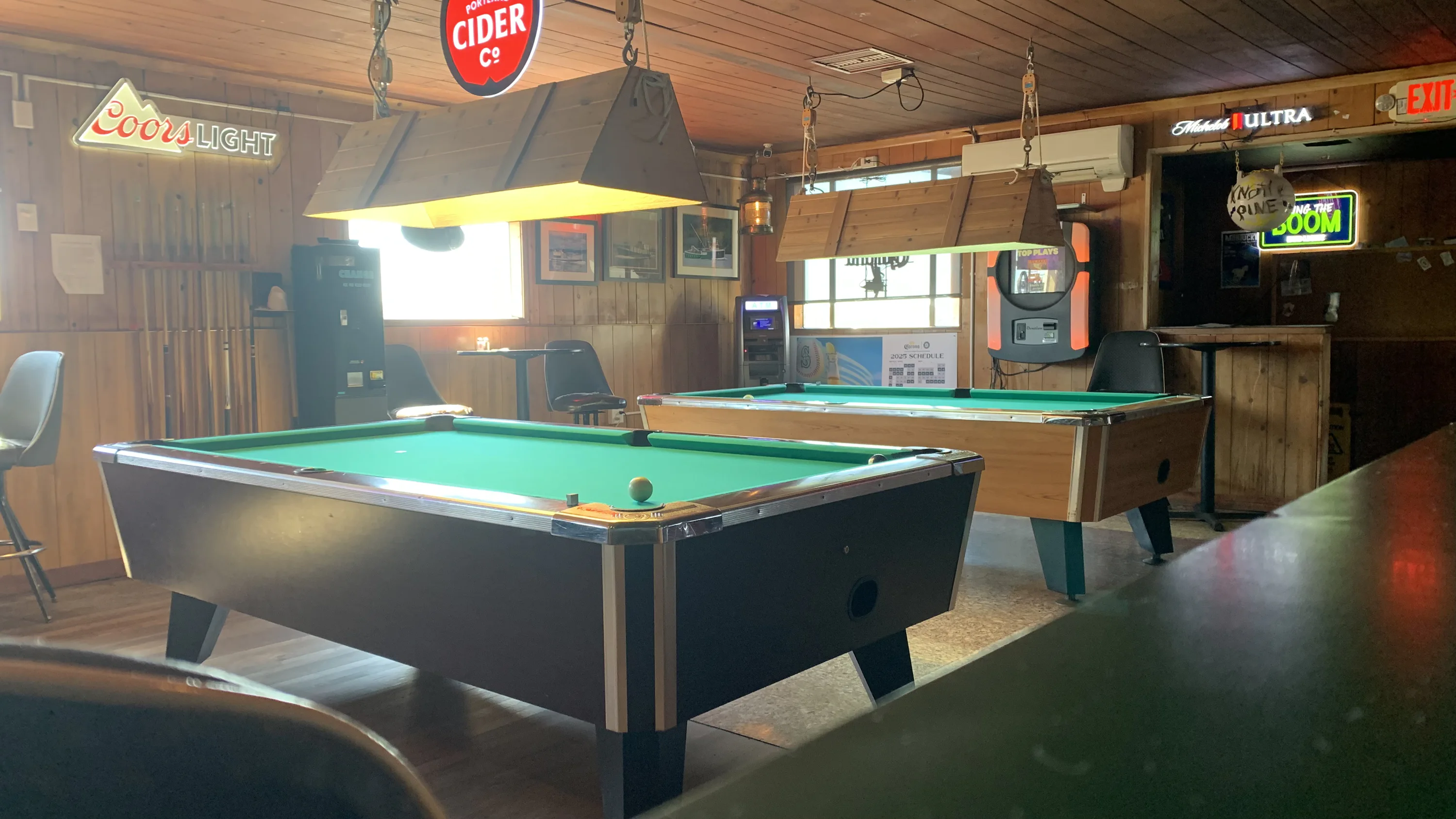 Dimly lit bar with two pool tables, wooden walls, bar stools, neon signs, and a jukebox. Cue sticks are displayed on the wall, with soft natural light filtering in.