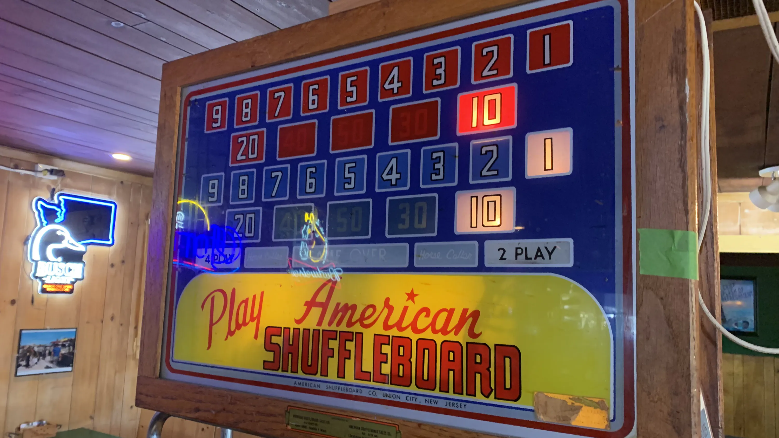 Vintage American Shuffleboard scoreboard with illuminated scores, mounted on a wooden panel in a rustic room. Neon signs visible in the background.