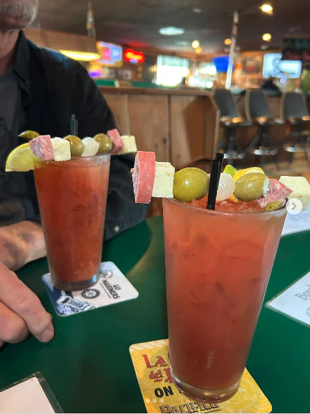 Two Bloody Mary cocktails with skewers of cheese, olives, meat, and lime wedges, served on a green table in a bar setting. A person sits nearby.