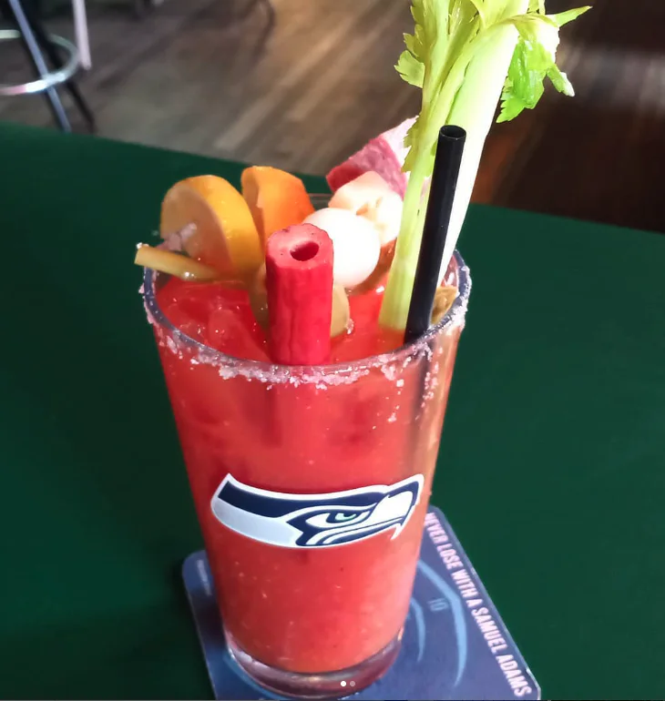 A Bloody Mary cocktail in a Seattle Seahawks glass, garnished with celery, lemon slices, pickles, meat, and a straw, placed on a bar counter with a blue coaster.