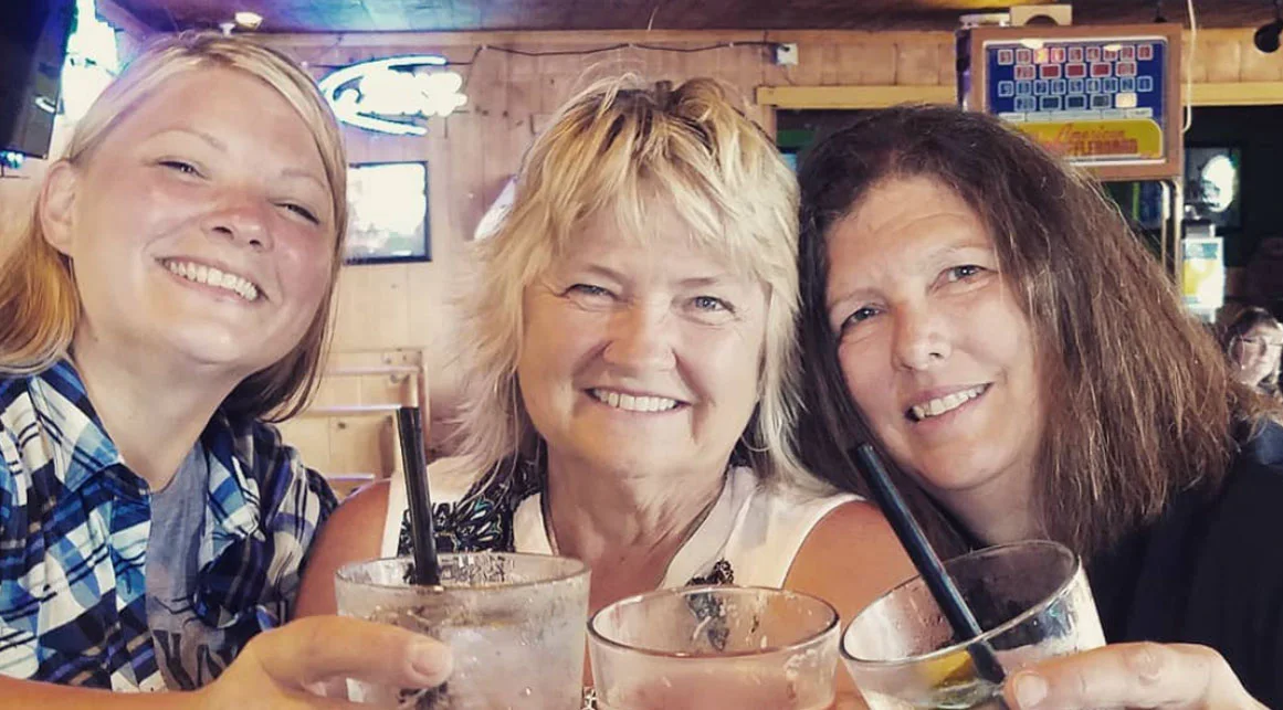 Three women smiling and holding drinks with straws in a cozy bar setting. Neon signs and wooden walls are visible in the background.