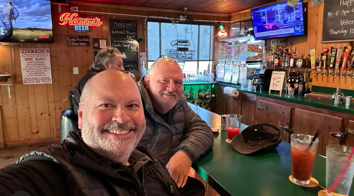 Two people smiling at a bar, seated with drinks on the counter. The bar has wooden walls, various signs, and two TVs mounted, one playing Netflix and the other showing news.