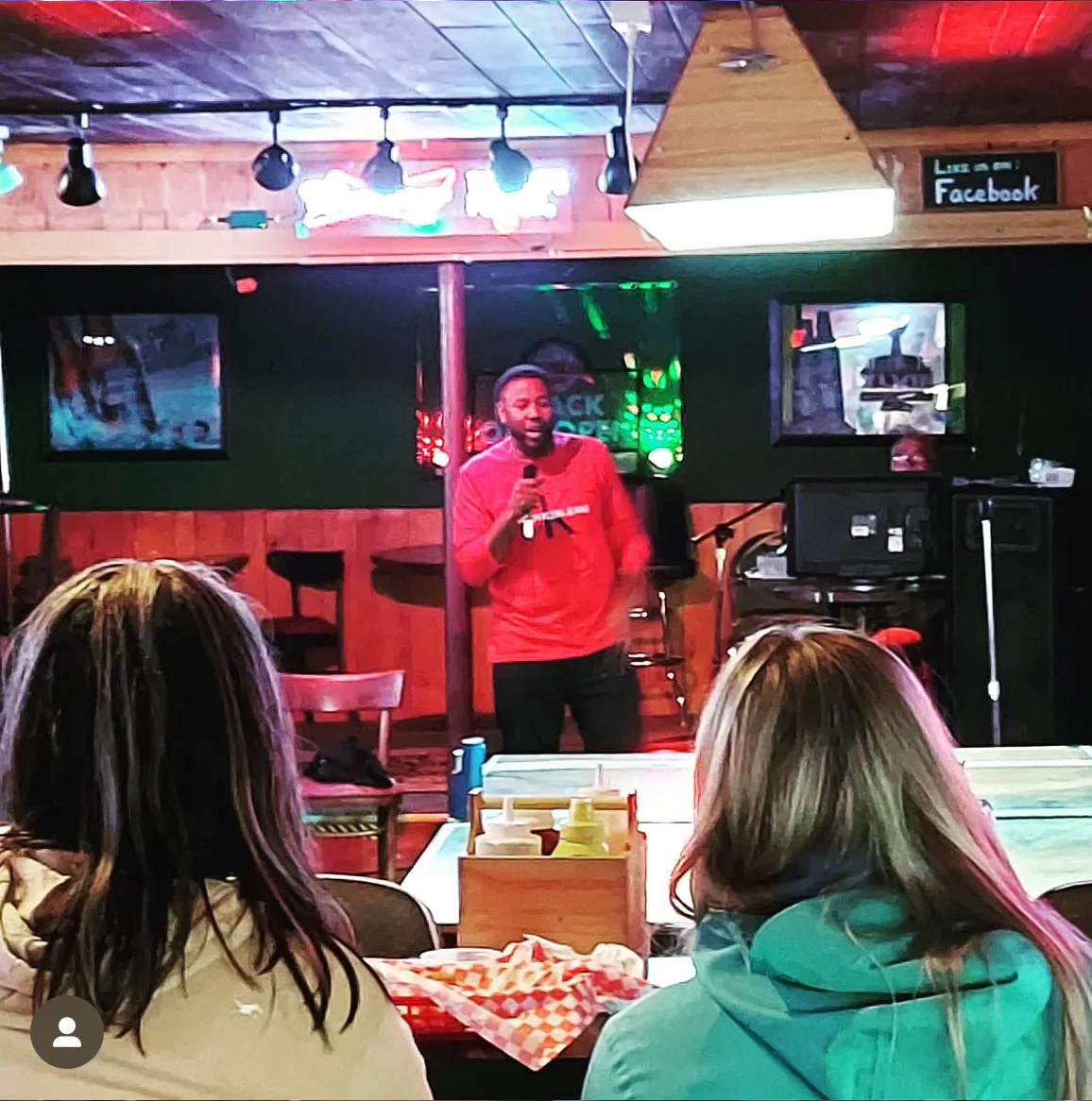 A person in a red shirt is performing on stage with a microphone in a cozy venue. Two audience members are seated in the foreground, watching attentively.