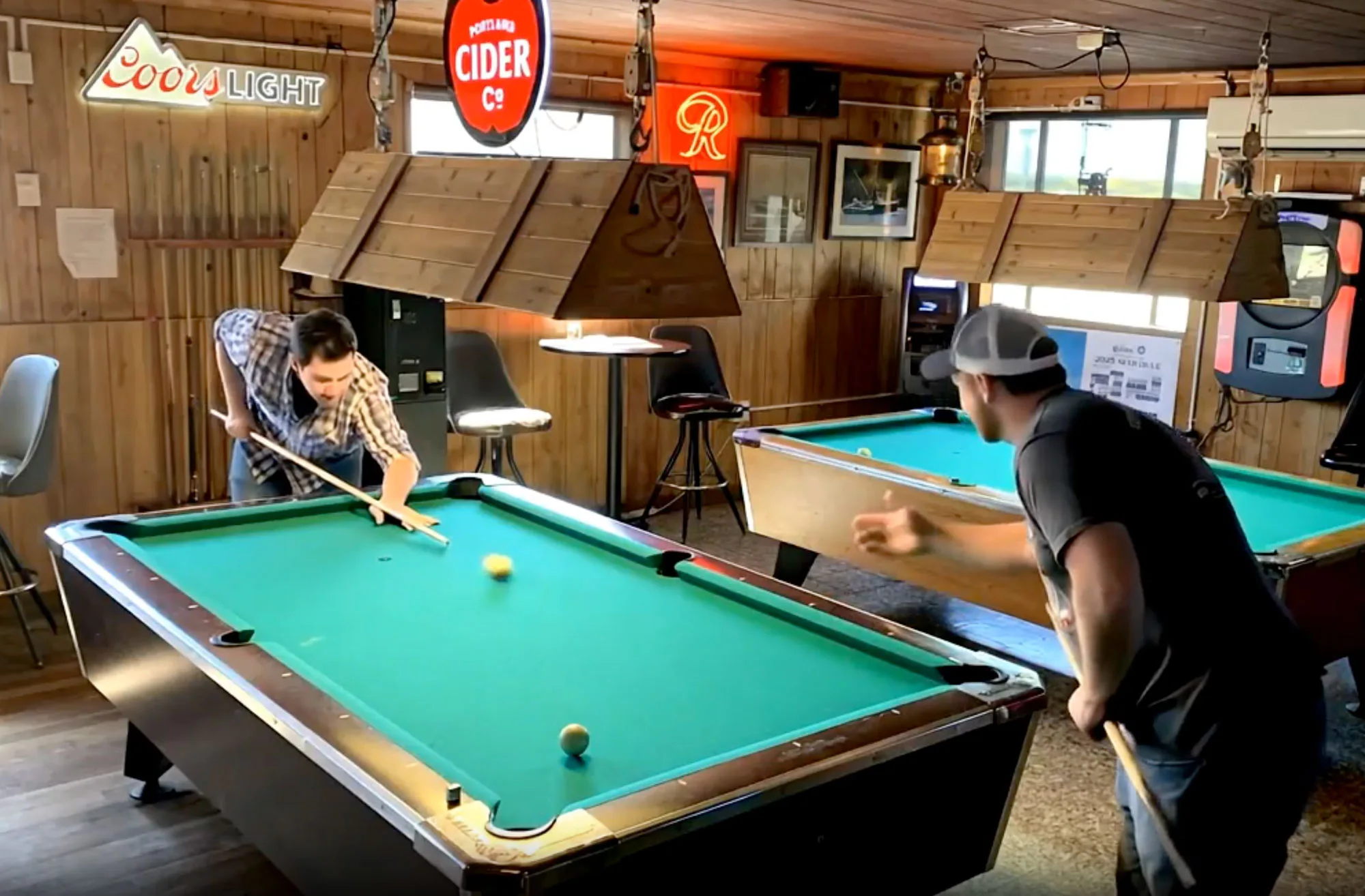 Two men playing pool in a bar with wood-paneled walls. Various neon signs, bar stools, and a jukebox are visible in the background.